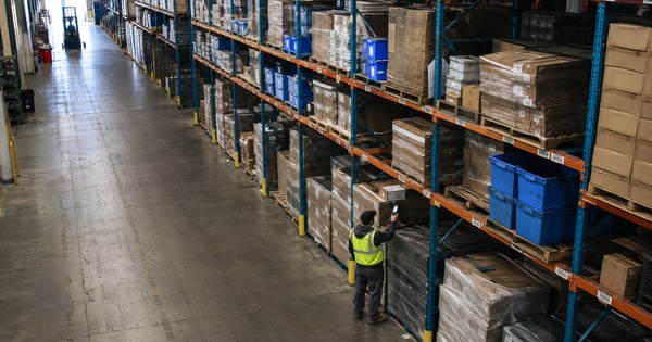 Warehouse employee using a handheld scanner to verify inventory along rows of pallet racks in a distribution center.