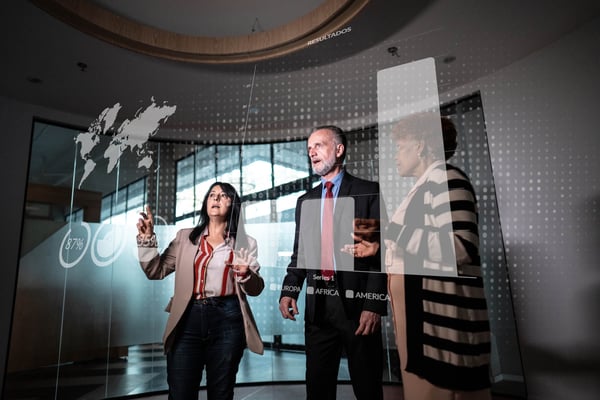 Three business professionals reviewing interactive data visualizations and global analytics displayed on a transparent digital screen in a modern office.